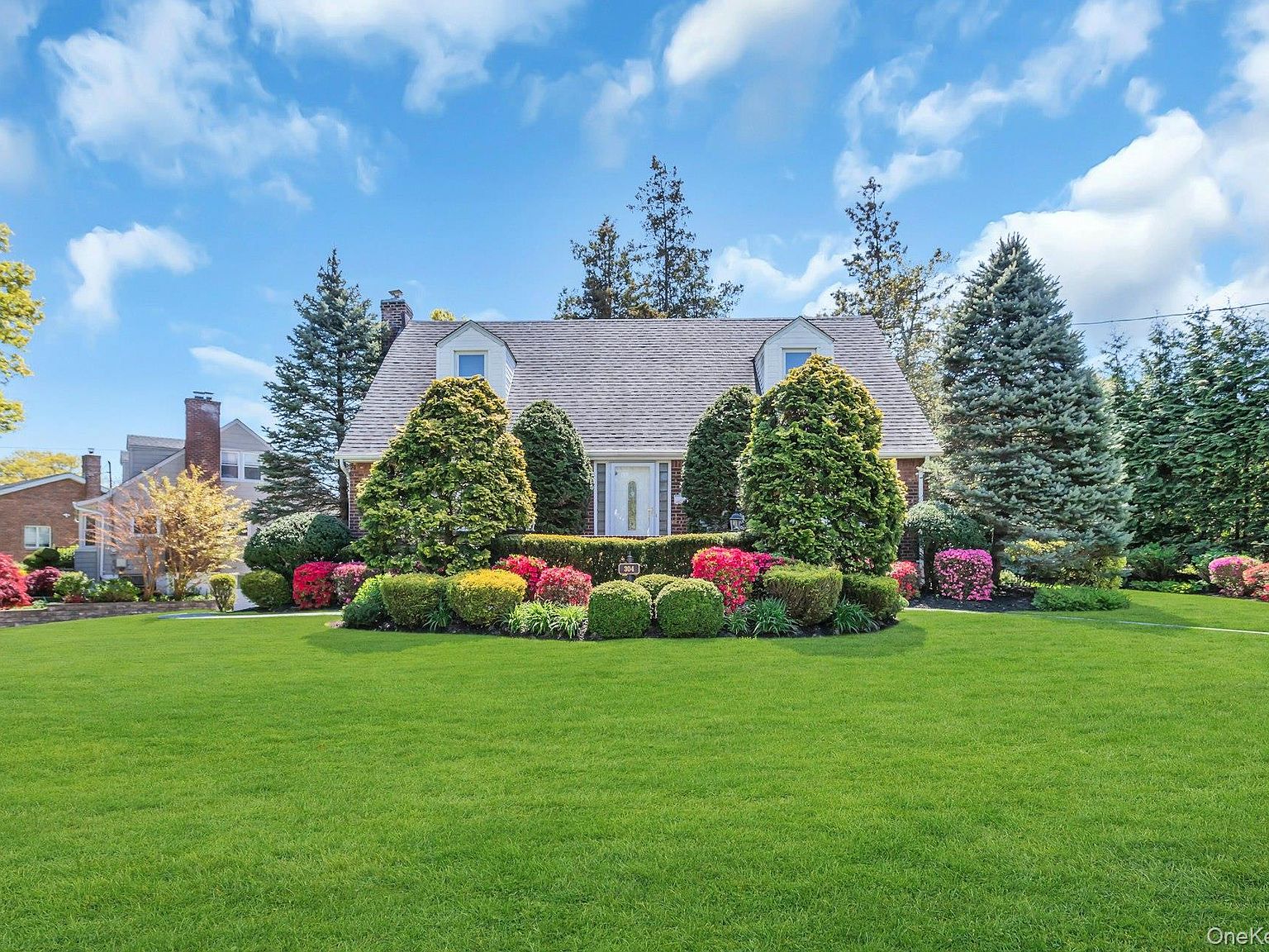 The brick Cape Cod facade of 304 Old Courthouse Road, fronted by manicured boxwoods and blooming azaleas under a bright spring sky